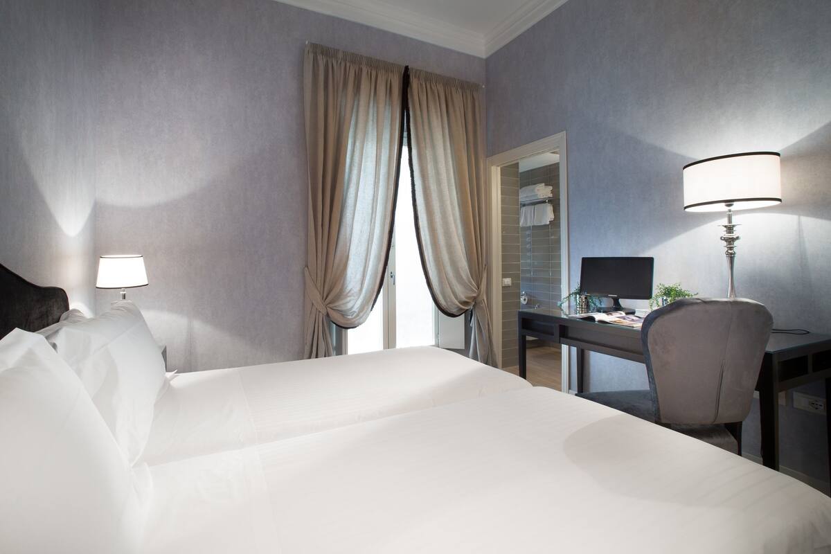 A hotel room with a bed, a desk, a chair, a lamp, and a window with curtains.