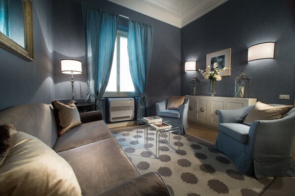A living room with a sofa, armchair, and a coffee table. There is a window with curtains, a lamp, and a picture on the wall. - San Giuliano Inn - Adults Only (Florence)