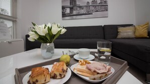 A dining table with a tray containing pastries, a pear, and a glass of water. A vase with white flowers is on the table, and a black and white cityscape painting is on the wall behind. - Harmony House (Bracknell)