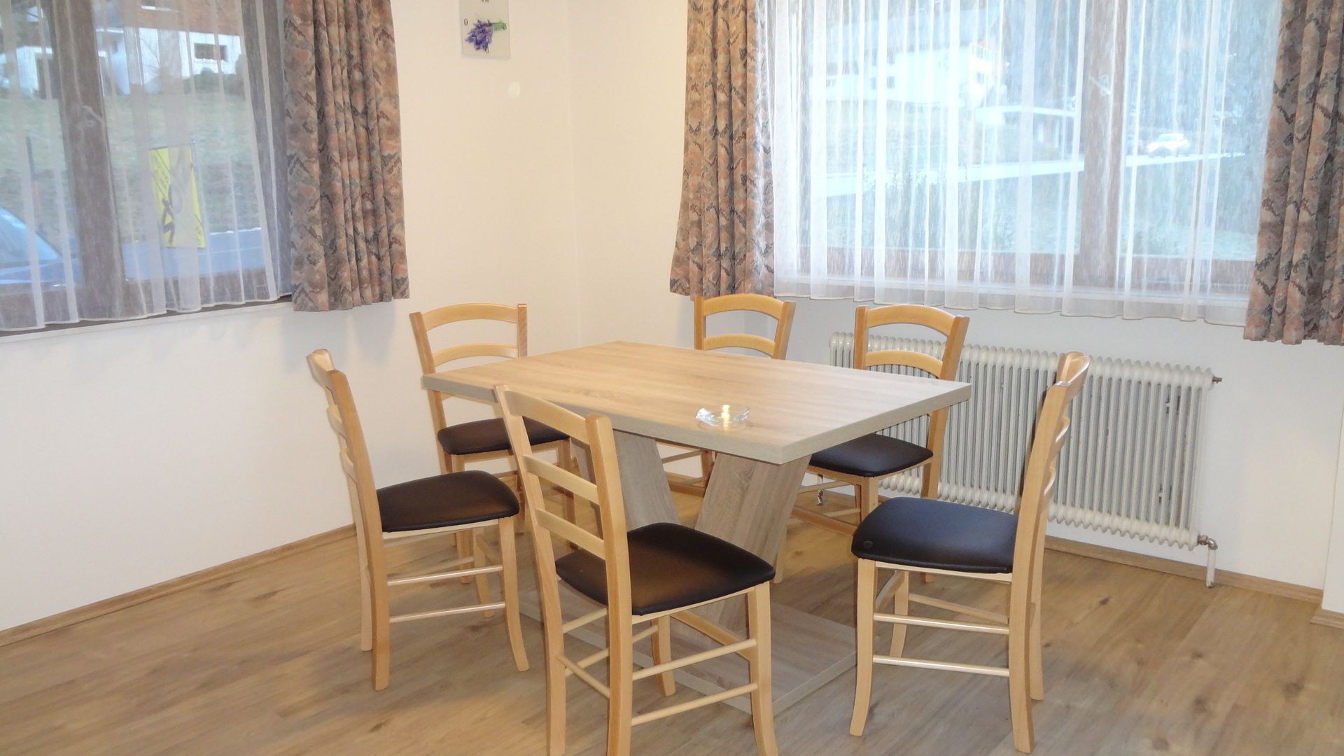 Een eetruimte met een houten tafel en zes stoelen, een radiator en gordijnen voor de ramen.