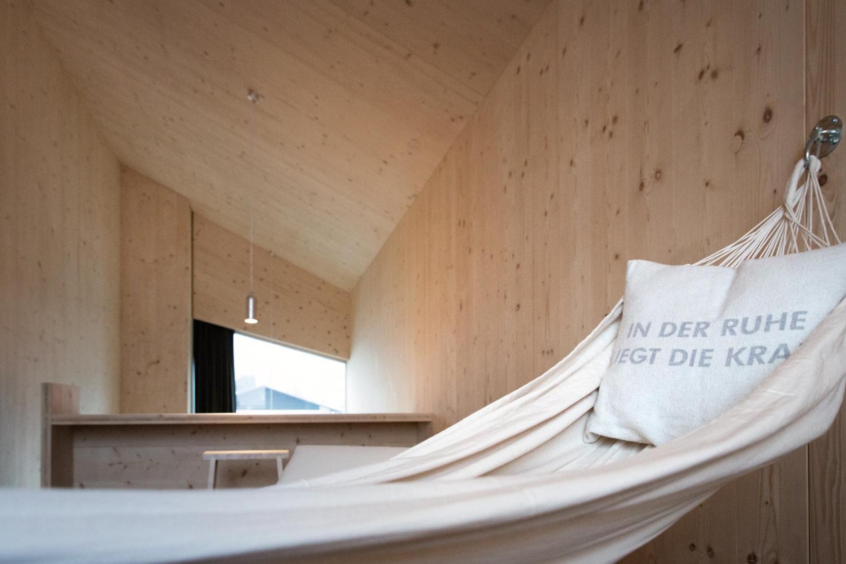 A hammock bed with a pillow that has text on it, in a wooden room with a sloped ceiling and a window.