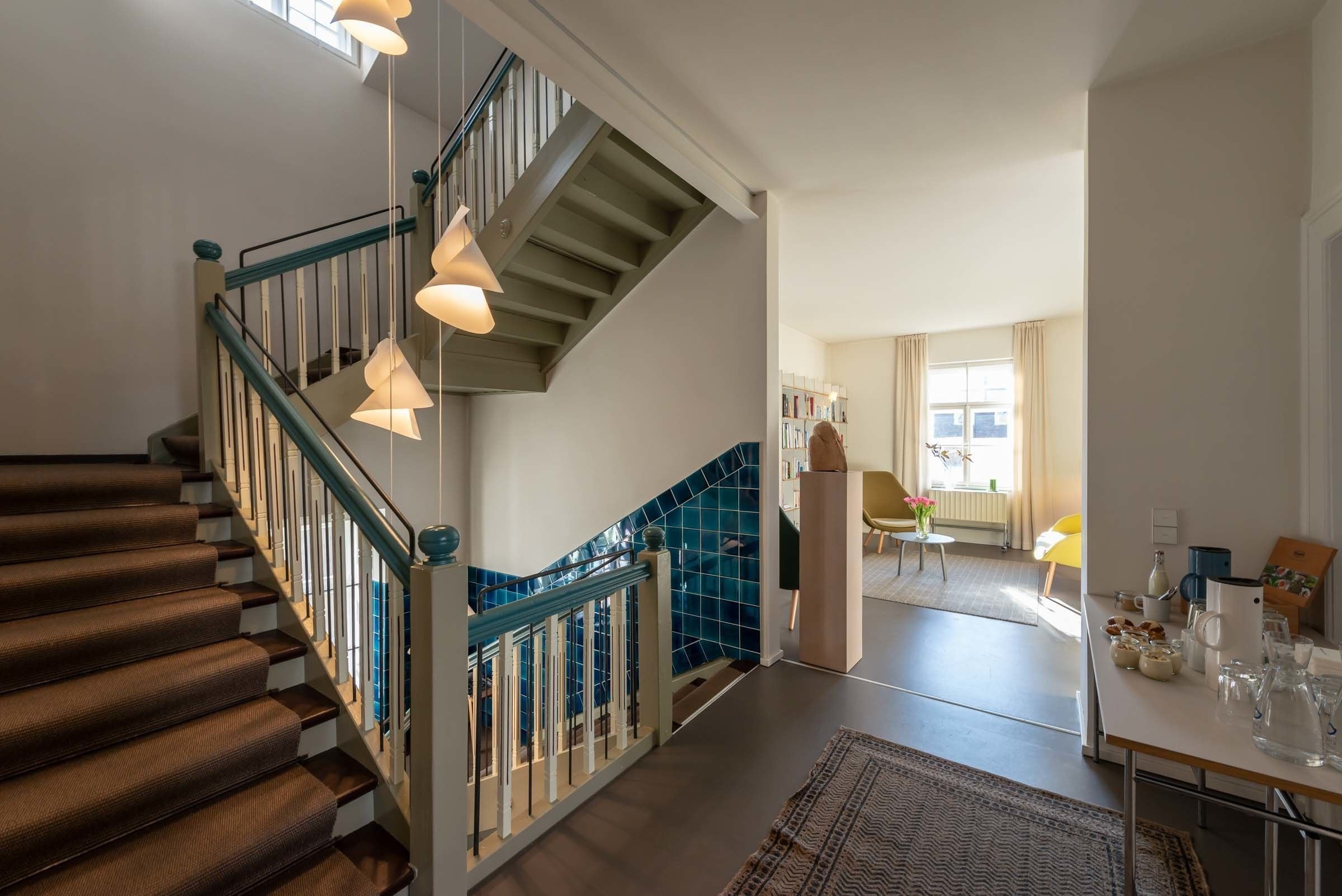 A staircase with wooden steps and blue railings, leading to a room with a tiled wall and a window with curtains.