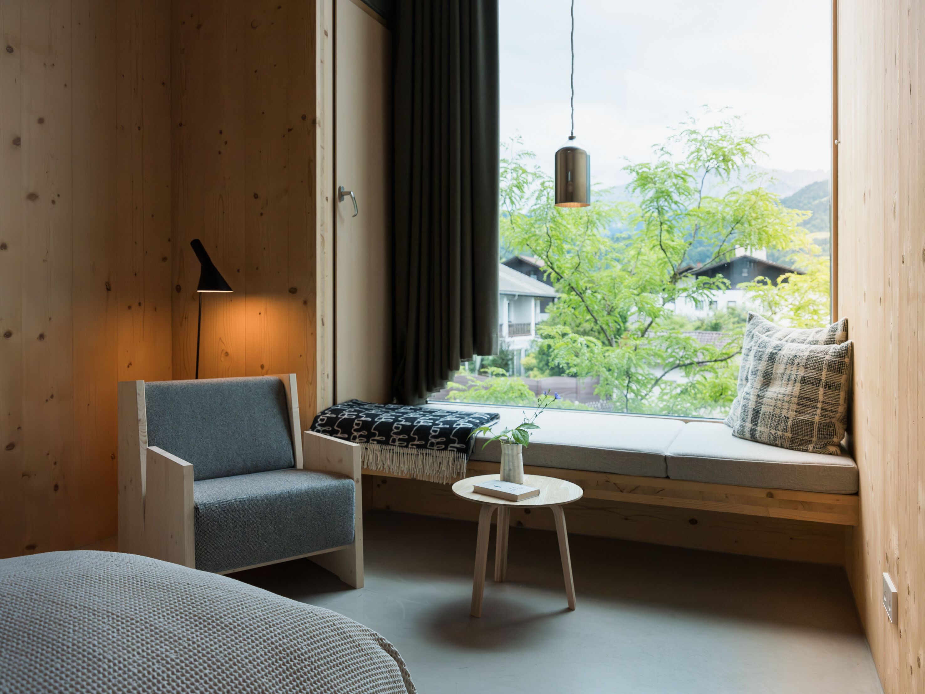 A cozy room with a wooden interior, a comfortable chair, a small table with a book, and a view of greenery and houses outside.