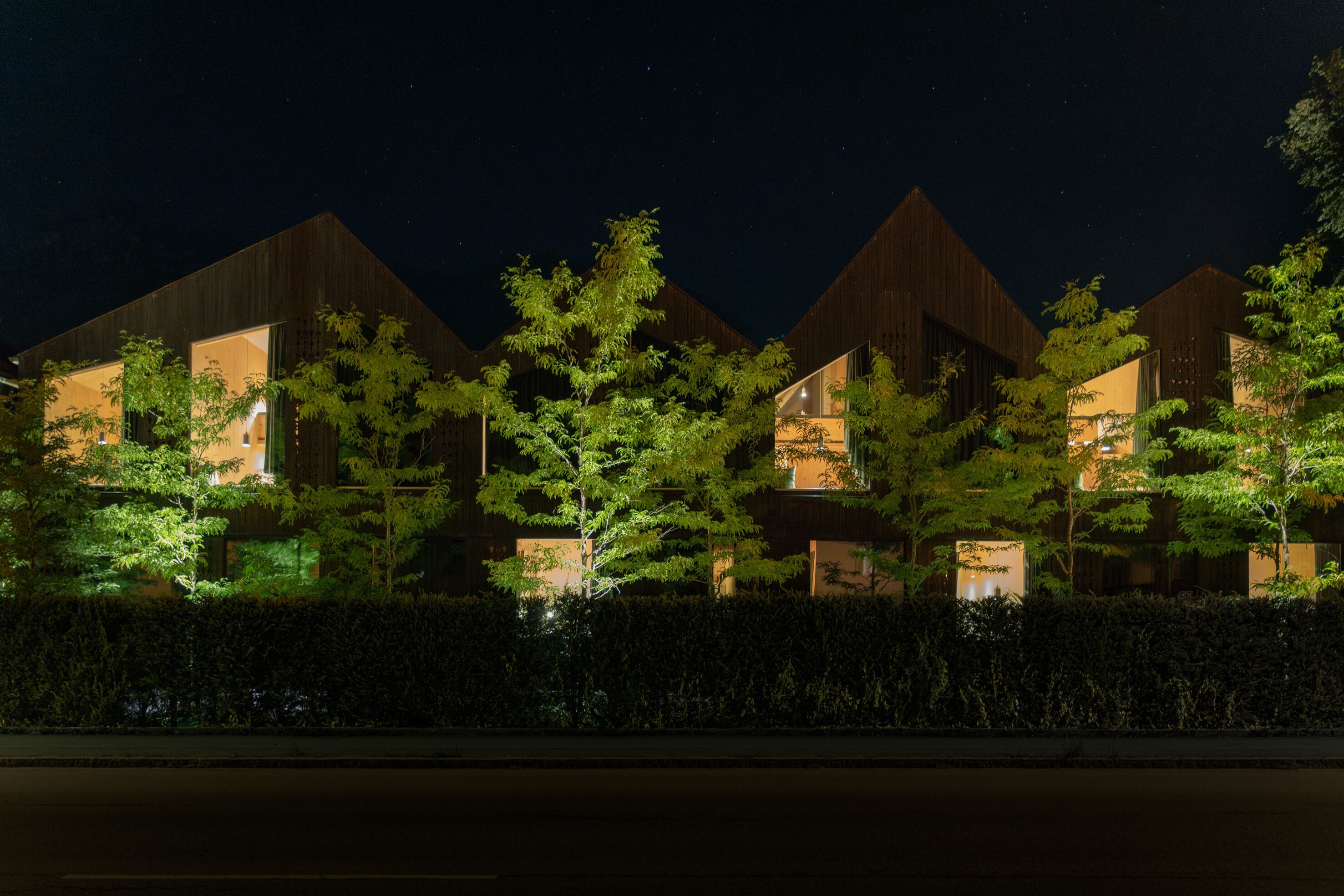 A row of modern, triangular-roofed buildings at night, illuminated by internal lighting, surrounded by trees and a hedge.