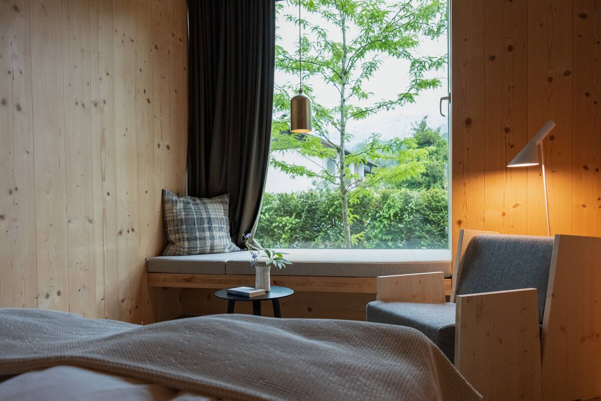 Ein gemütliches Schlafzimmer mit einem großen Bett, einem kleinen Tisch mit Büchern, einem Stuhl und einer Sitzbank am Fenster mit Blick auf Grünfläche.