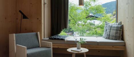 A cozy room with a wooden floor, a sofa with cushions, a small round table, and a window with a view of greenery and buildings.
