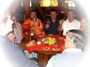 En grupp människor sitter runt ett träbord med en röd duk på, de håller i varsin drink och en flagga.