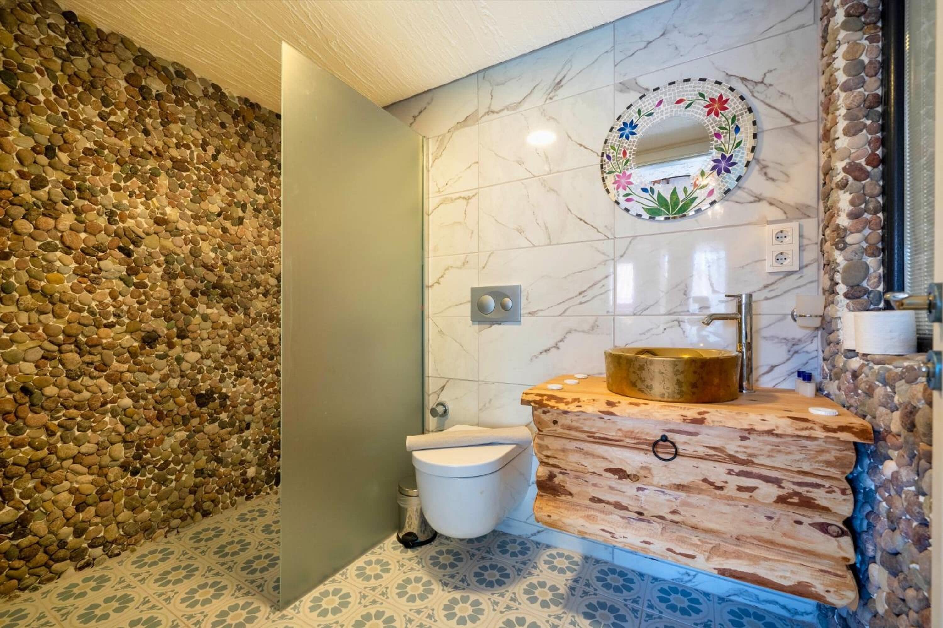 A bathroom with a stone wall, a marble-tiled floor, a wooden vanity with a gold sink, and a decorative mirror.