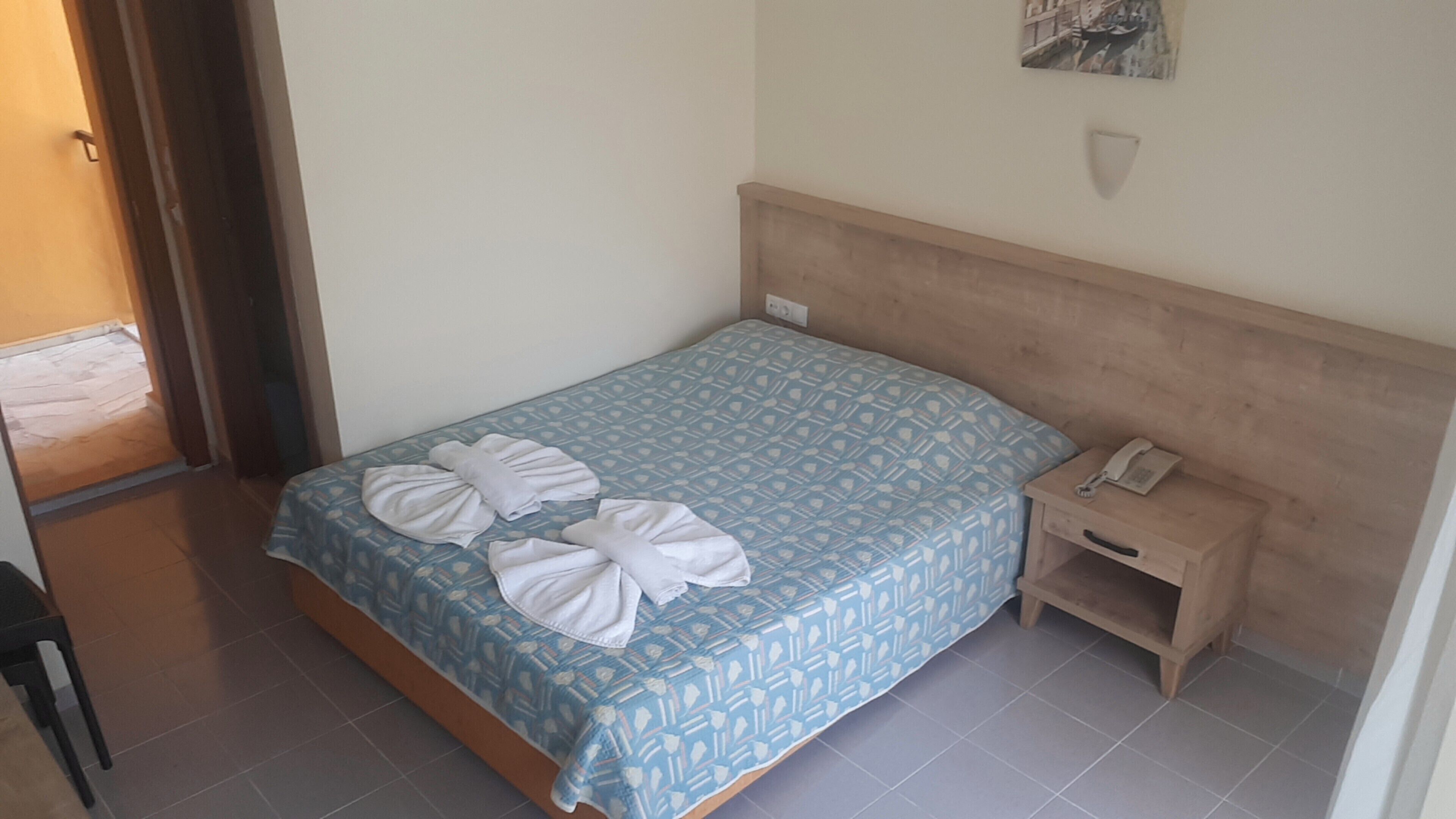 Una cama pequeña y bien hecha, con una colcha azul estampada y dos toallas dobladas encima.