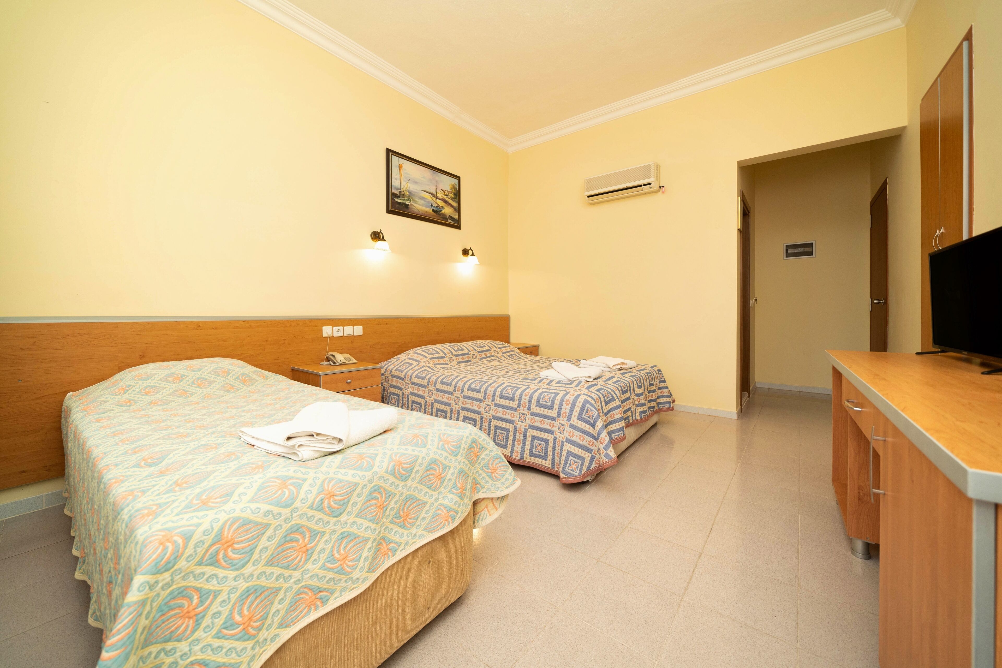 Habitación de hotel con dos camas, cabecero de madera, mesita de noche con lámpara, televisor y unidad de aire acondicionado en la pared.