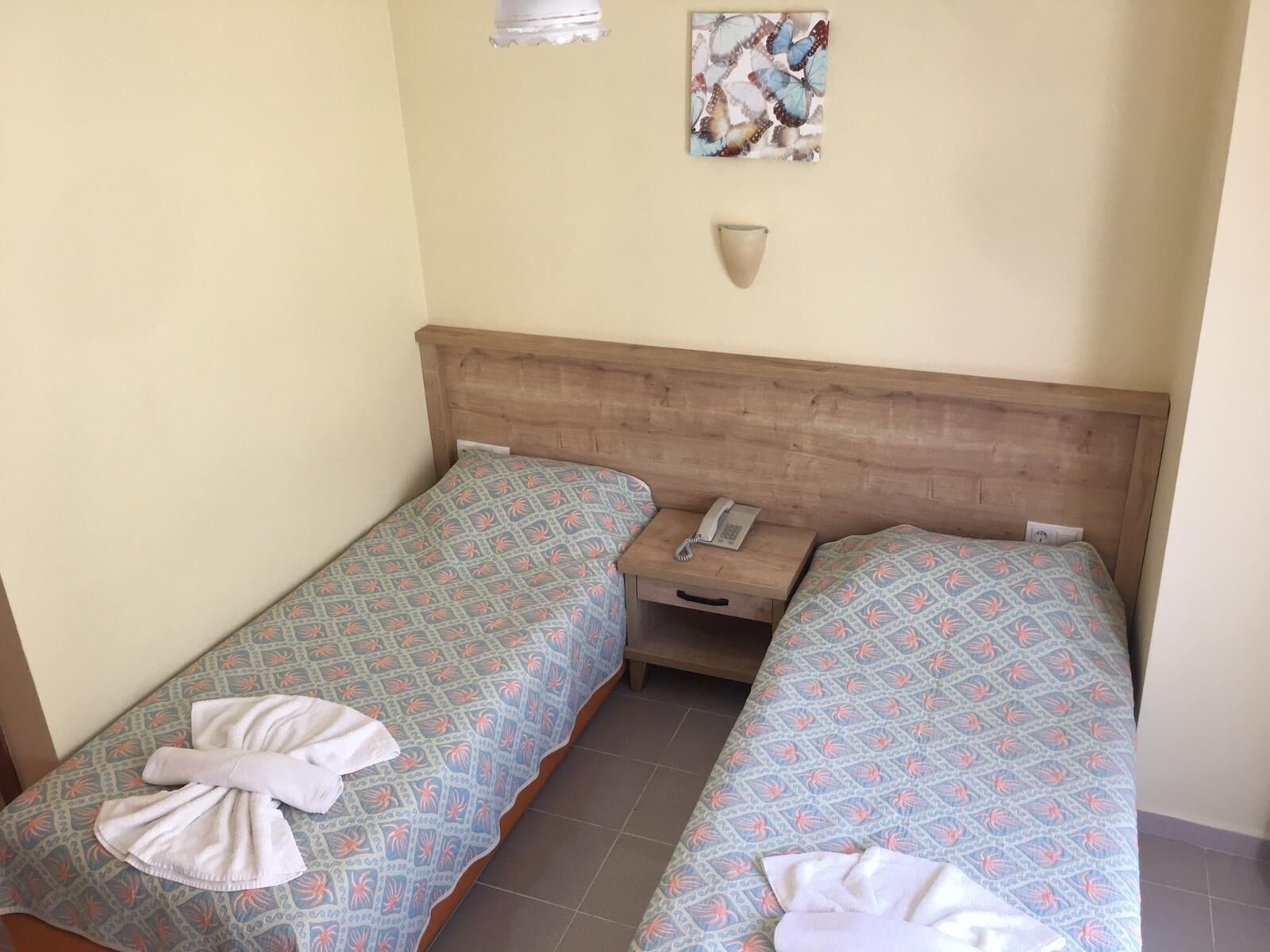 A double room with two beds, each with patterned bedding and towels. There is a bedside table with a phone and a wall-mounted light.
