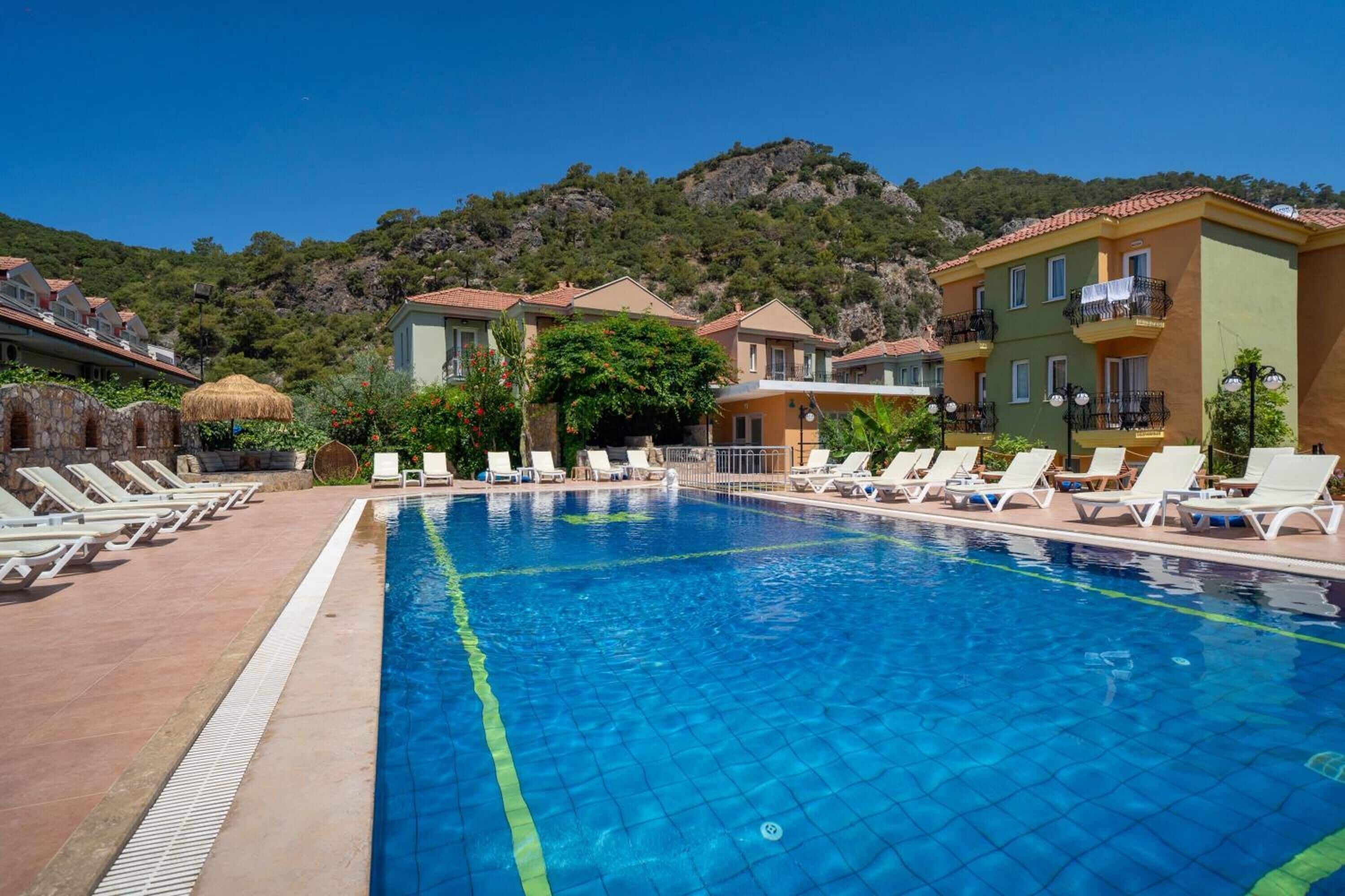 Uma piscina com espreguiçadeiras, montanhas ao fundo e edifícios com varandas.