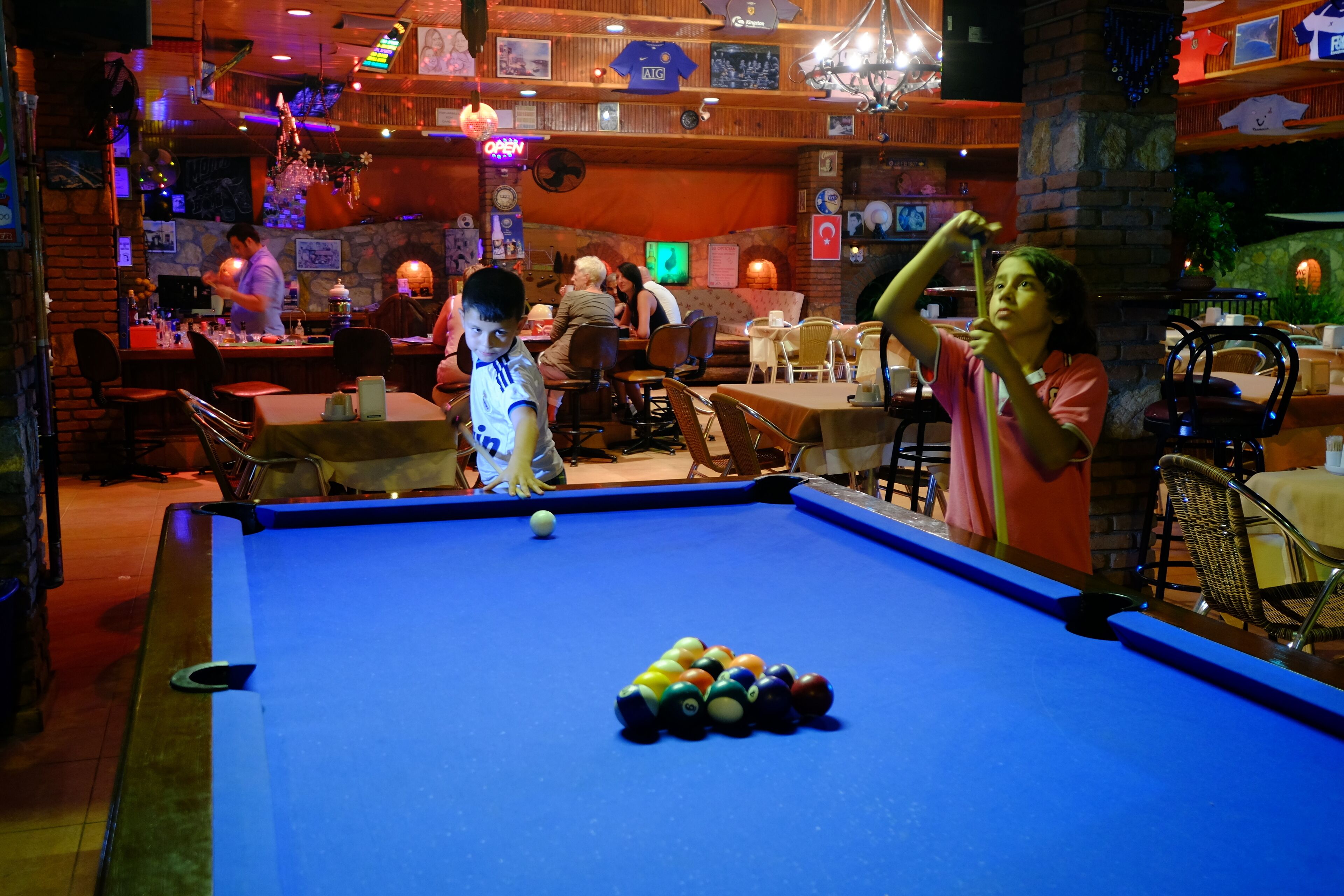 A pool table with balls, surrounded by tables and chairs in a dimly lit indoor setting.