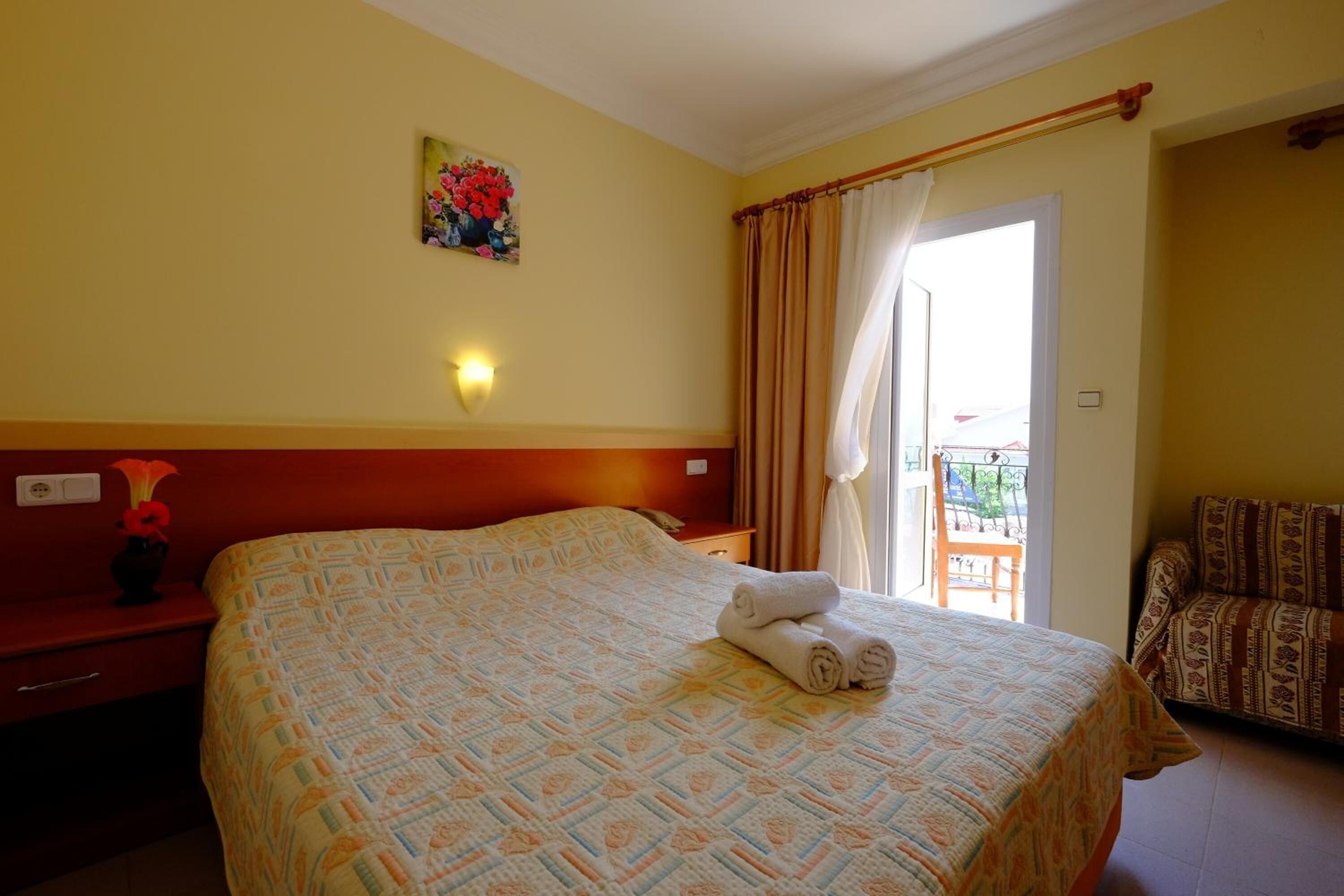 A hotel room with a large bed, a painting on the wall, a nightstand with a vase, and a view of the outside balcony.