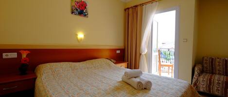 A hotel room with a large bed, a painting on the wall, a nightstand with a vase, and a view of the outside balcony.