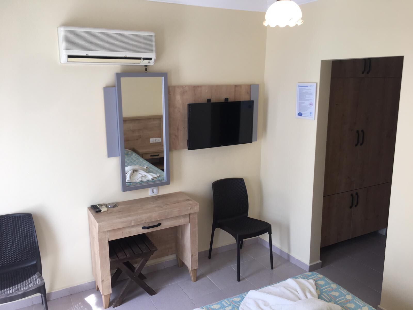 A room with a wooden desk, a chair, a bed, a wardrobe, a television, and an air conditioning unit.