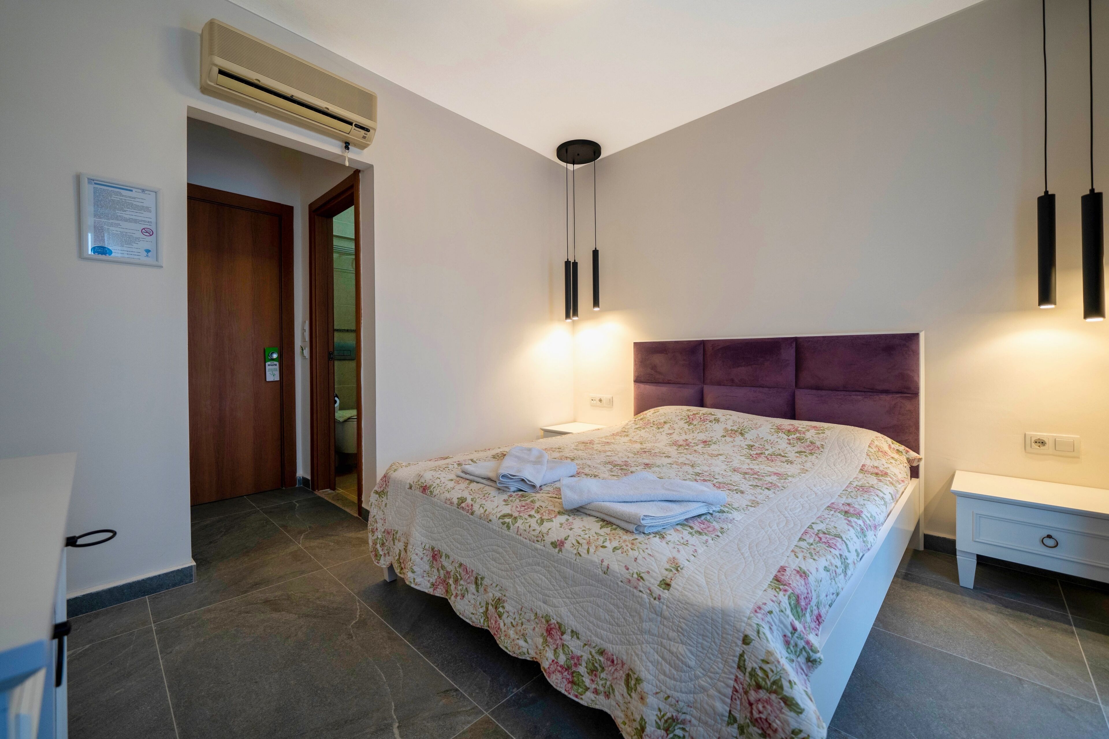 A bedroom with a floral bedspread, a headboard, a bedside table, and a wall-mounted air conditioner.