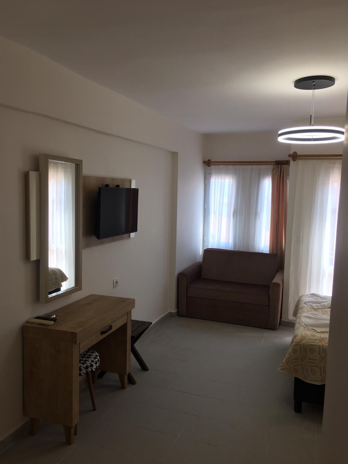 Une chambre d’hôtel compacte comprenant un bureau en bois, un canapé, un lit, un téléviseur fixé au mur et un luminaire au plafond.
