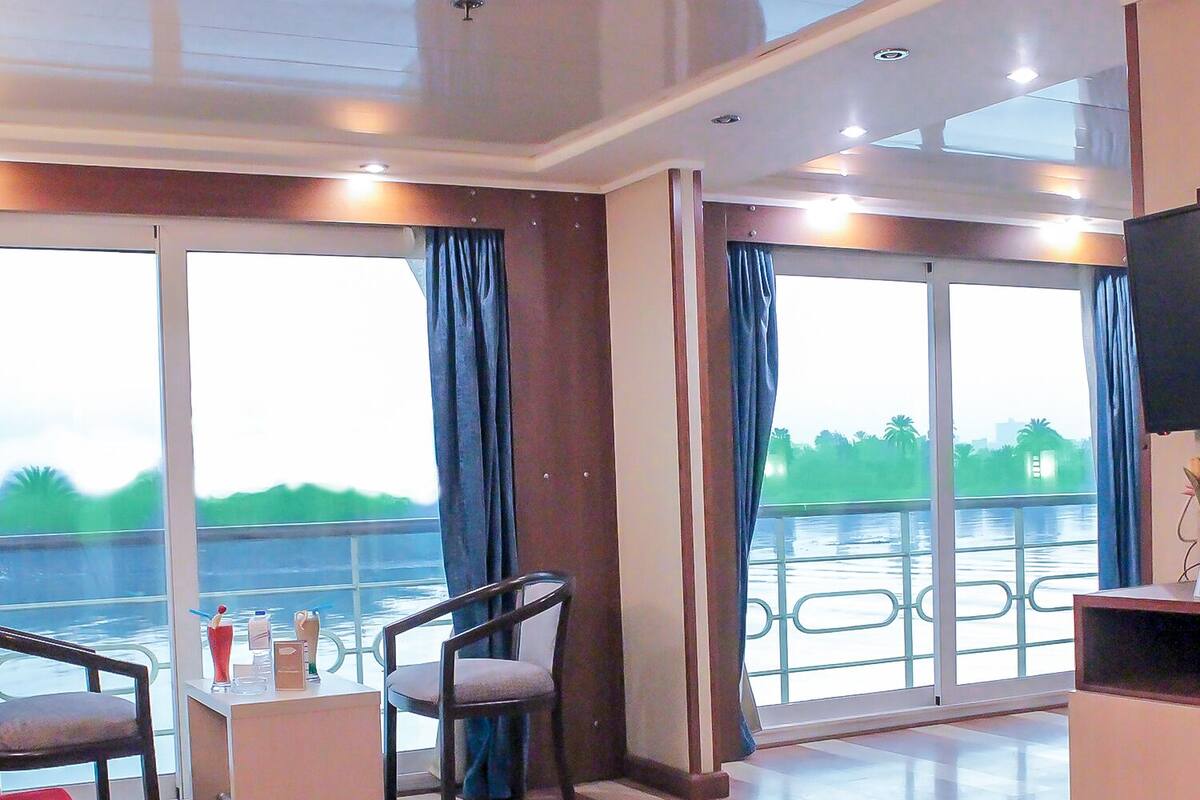 A modern hotel room with a large window offering a view of a body of water and greenery. There is a seating area with a red sofa and a small table with a drink and a vase.