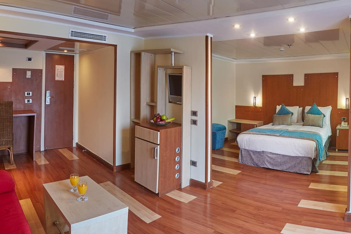 A modern hotel room with a wooden floor, a bed with a blue and white bedspread, a desk with a mirror, and a view of the water through the window.
