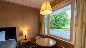A hotel room with a bed, a table with chairs, a lamp, and a window with a view of greenery and a parking lot.