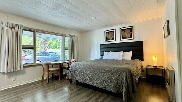 A bedroom with a bed, a desk, and a chair. There are two windows with curtains, a ceiling with recessed lighting, and two framed pictures on the wall. - Mount Coolidge Motel (Lincoln)