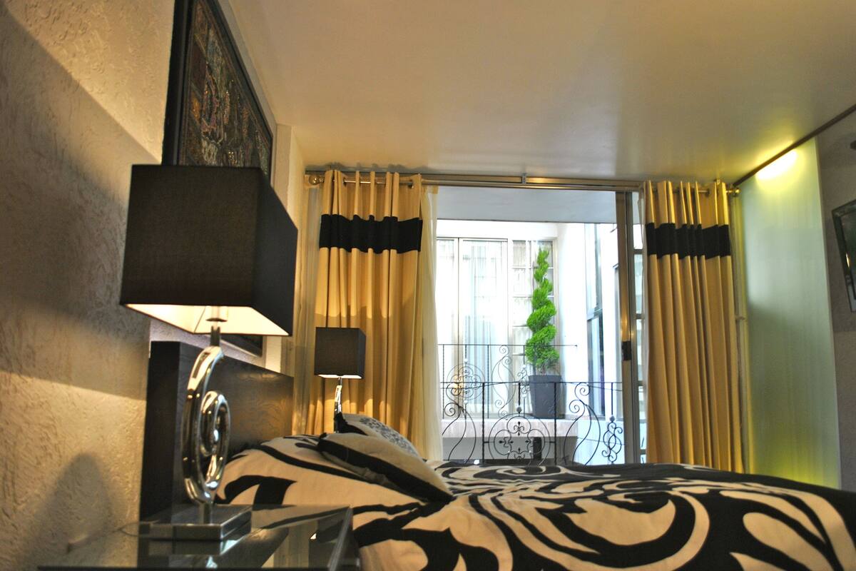 A hotel room with a bed, a nightstand, a lamp, a window with curtains, and a view of a balcony with a plant.