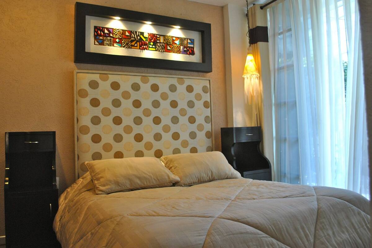 A hotel room with a polka dot headboard, a bed with beige bedding, a framed artwork above the headboard, a bedside table with a lamp, and a window with sheer curtains.