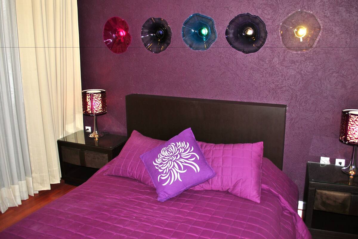 A bedroom with a bed, purple bedding, two bedside tables with lamps, and decorative wall lights.