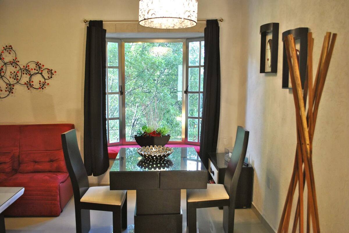 A dining area with a glass table, two chairs, a red sofa, a window with curtains, and a decorative wall piece.