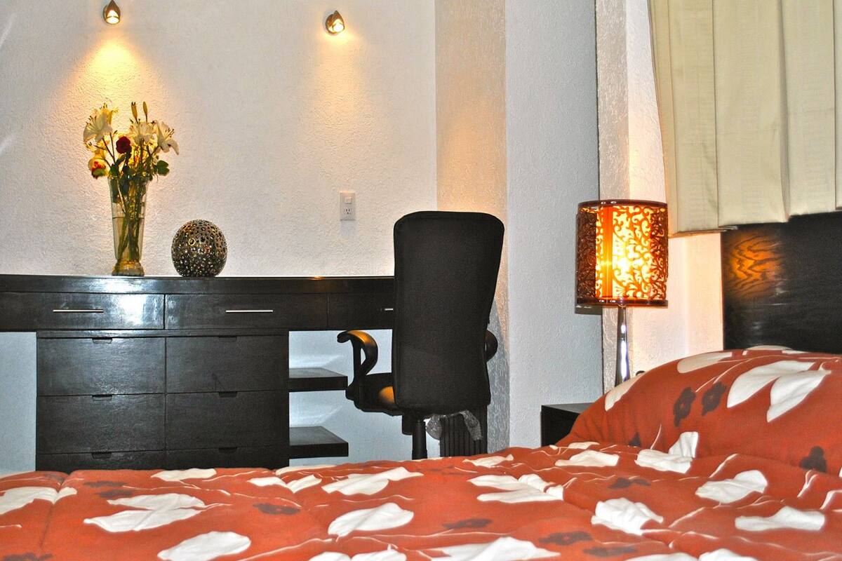 A hotel room with a bed, a desk with drawers, a chair, a lamp, and a vase with flowers.