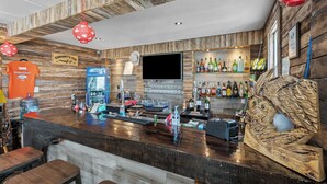 A bar area with wooden walls, a mounted TV, and a variety of bottles and glasses on shelves.