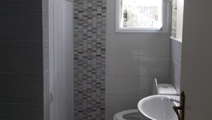 A bathroom with a tiled shower wall, a toilet, a sink, and a window providing natural light. - Iris Villa (Lefkada)