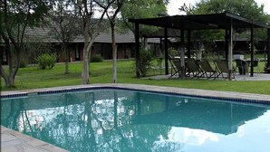 Una piscina rectangular con borde de azulejos, rodeada por una terraza de piedra. Se observan sillas de descanso y un área de estar cubierta al fondo.