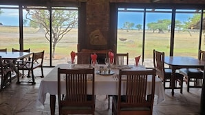 Un comedor con mesas y sillas, ventanas amplias y vistas a la sabana.