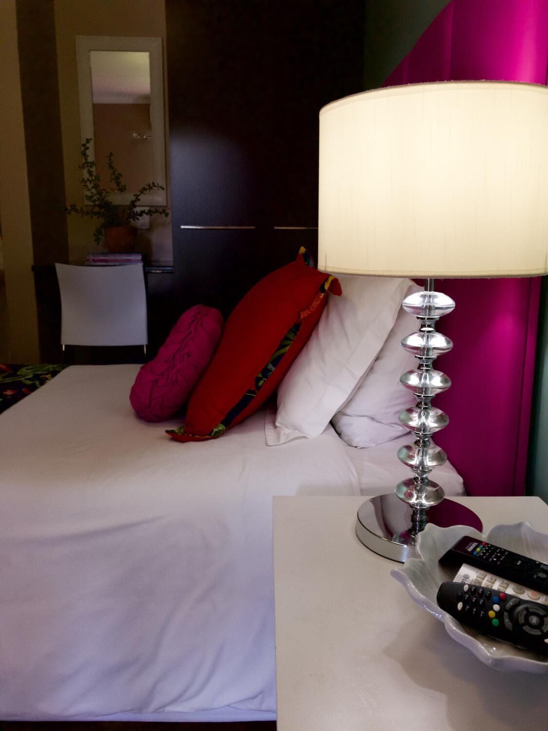 Habitación de hotel con cama, mesita de noche con lámpara y controles remotos, una silla y planta en maceta.