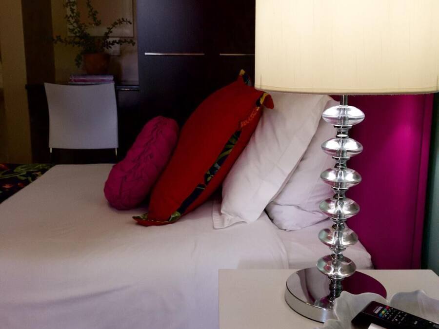 Habitación de hotel con cama, mesita de noche con lámpara y controles remotos, una silla y planta en maceta.