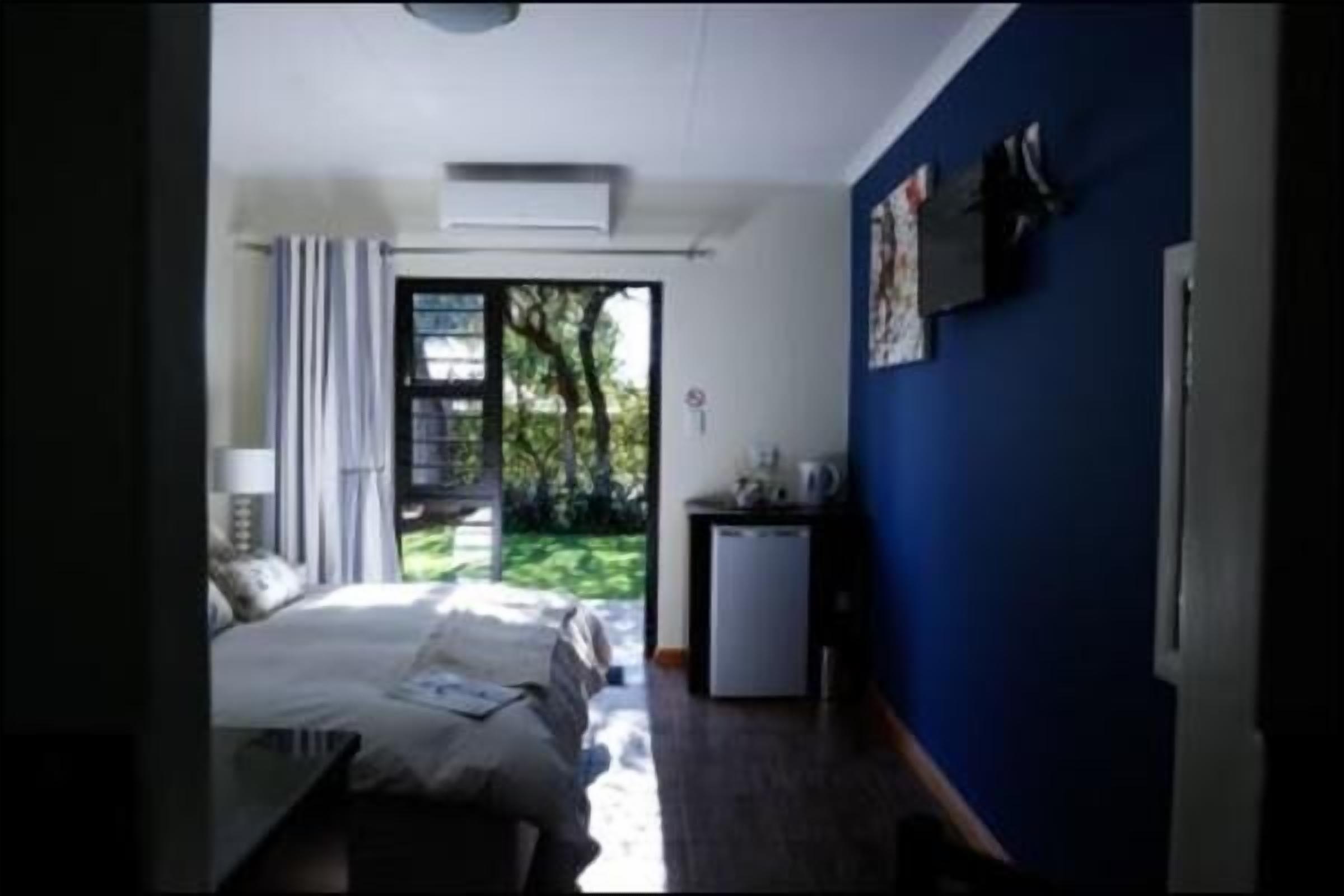 A bedroom with a bed, a desk, a chair, a small refrigerator, and a window with a view of greenery.