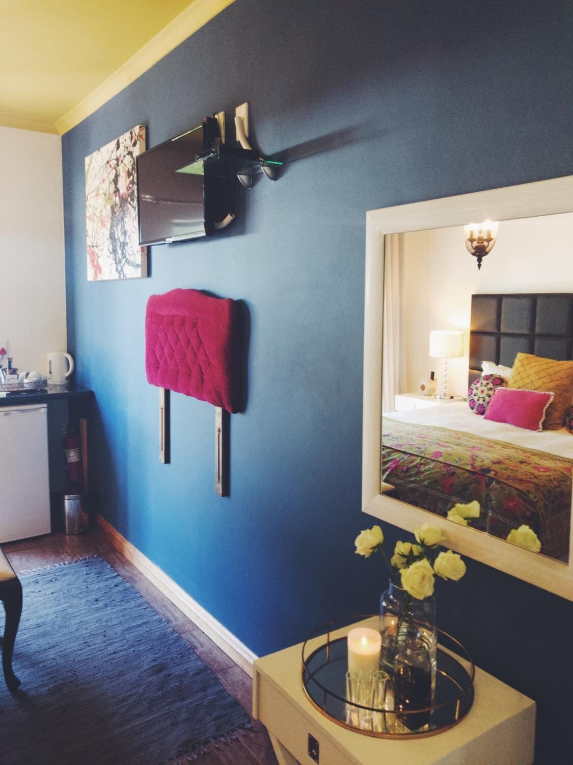 A bedroom with a bed, a bedside table with a vase, a mirror reflecting the room, and a wall-mounted TV.