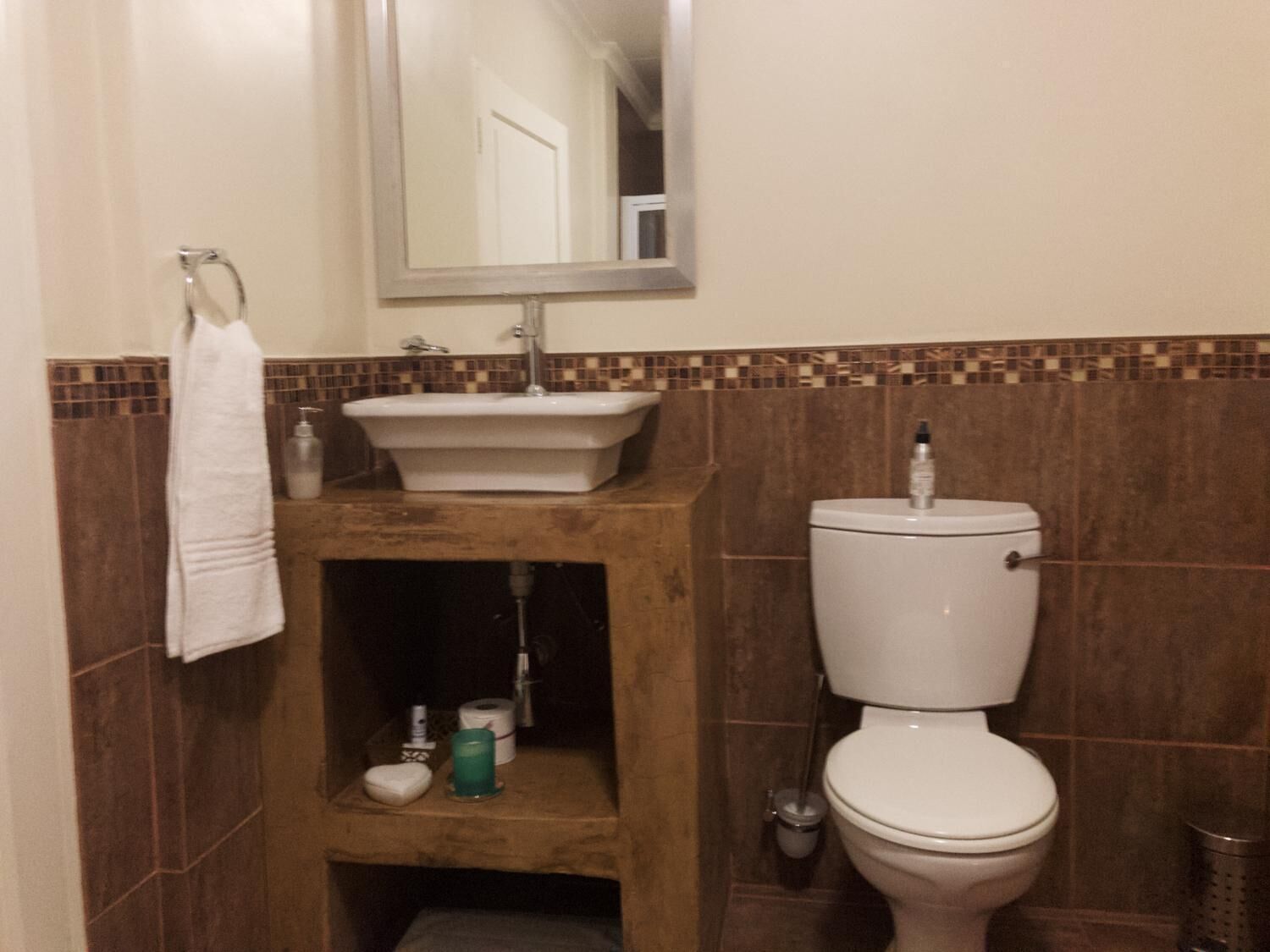 A bathroom with a white sink, a toilet, and a mirror above the sink. There is a towel rack on the left and a shelf below the sink with toiletries.