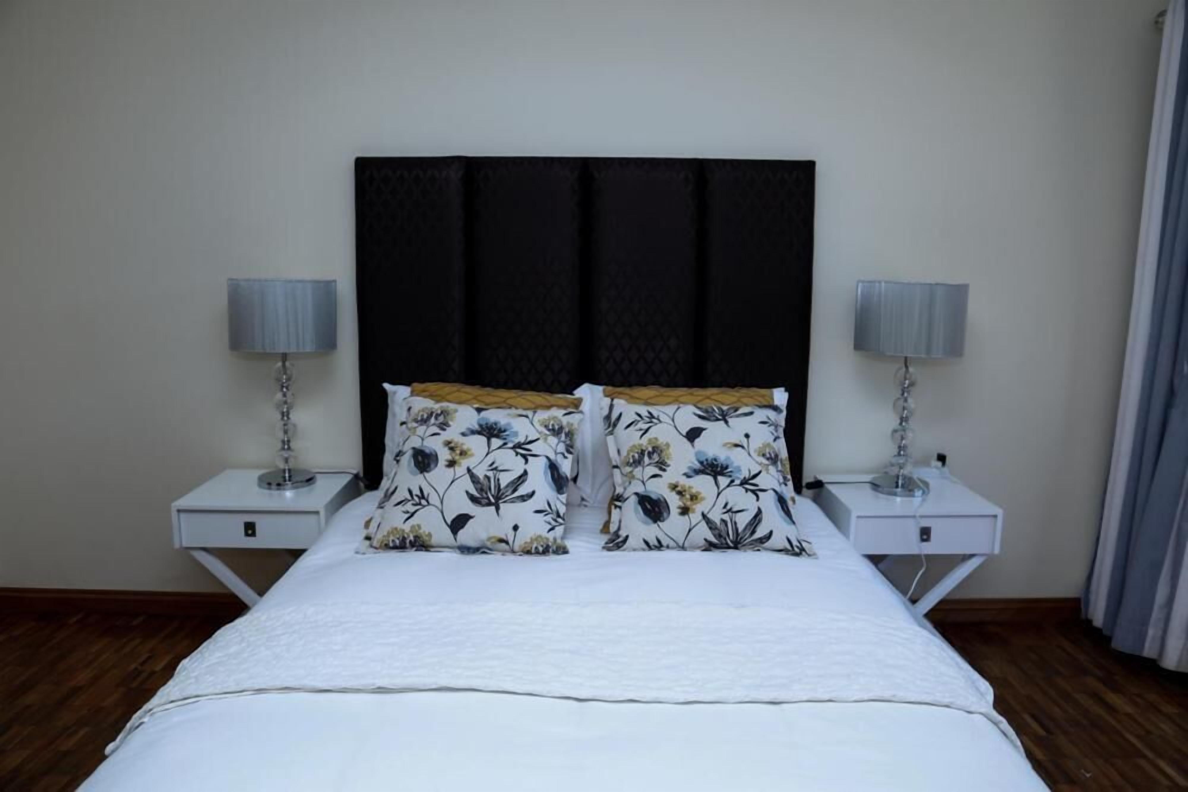 Cama bem arrumada com edredom branco e almofadas estampadas, com duas mesas de cabeceira com luminárias, num quarto com soalho de madeira e paredes lisas.