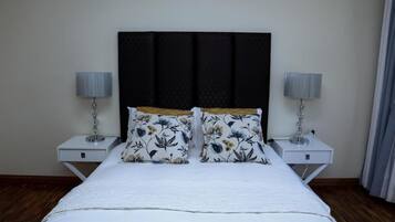 Cama bem arrumada com edredom branco e almofadas estampadas, com duas mesas de cabeceira com luminárias, num quarto com soalho de madeira e paredes lisas.