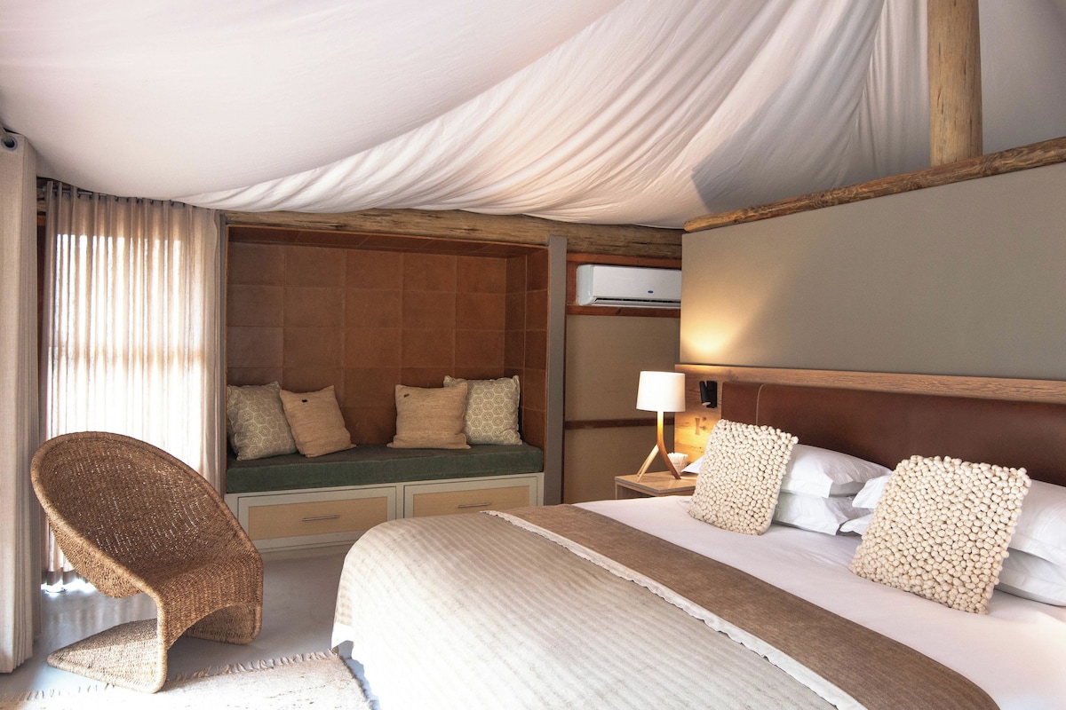 A hotel room with a bed, a seating area with cushions, a wicker chair, a nightstand with a lamp, and an air conditioning unit.