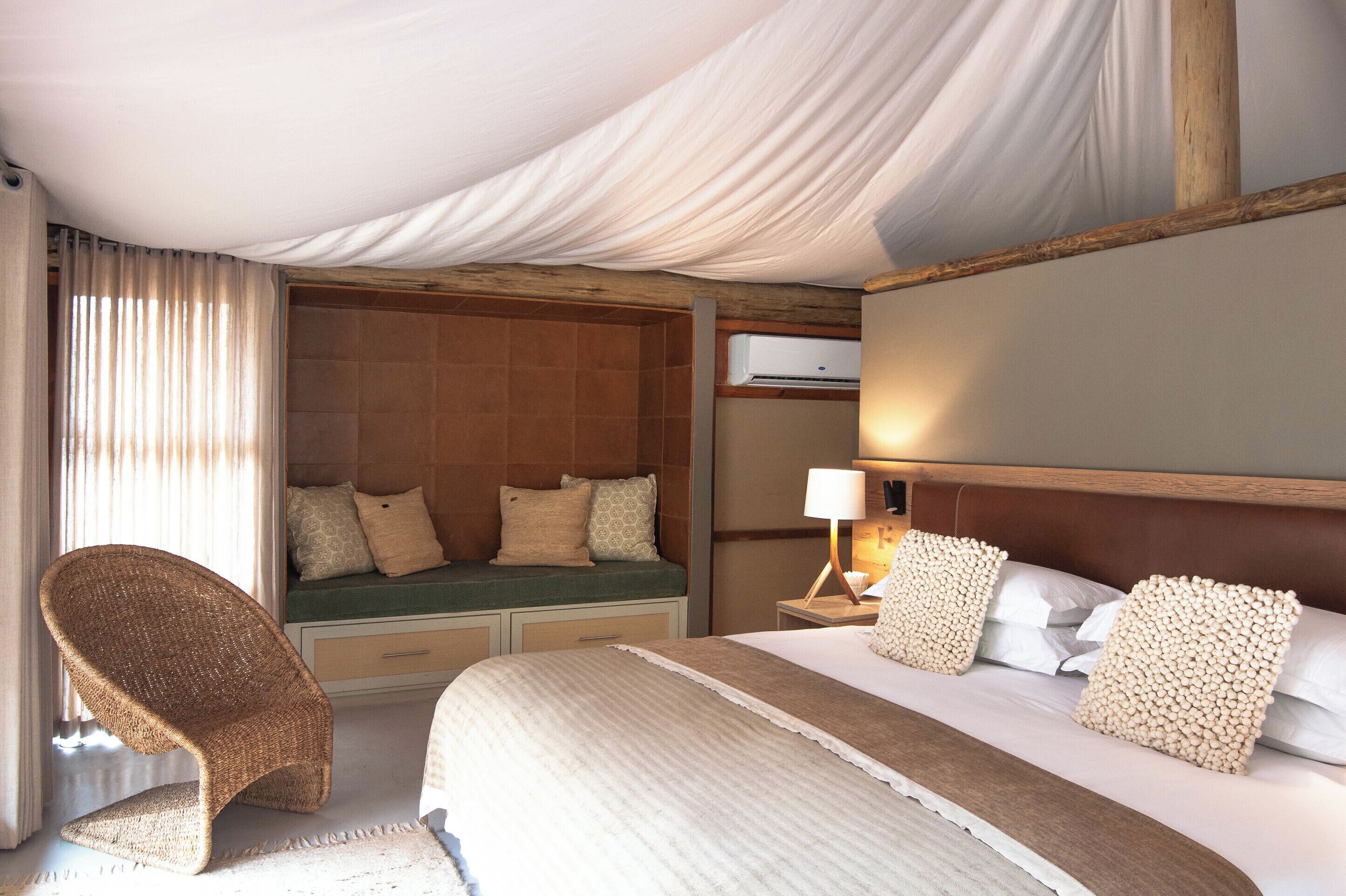 Een hotelkamer met een bed, een zithoek met kussens, een stoel, een nachtkastje met een lamp en een airconditioning.