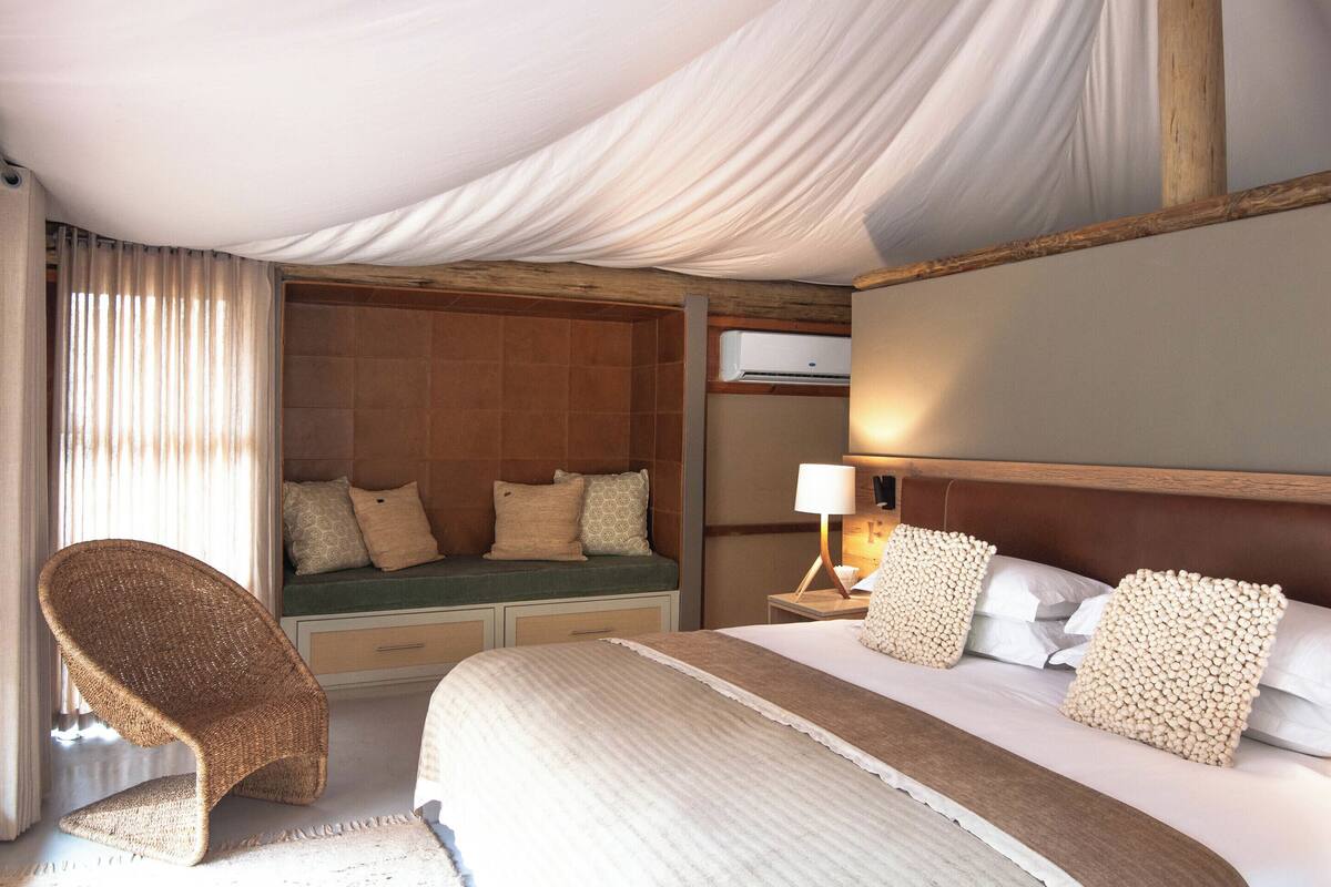 Ein Hotelzimmer mit einem Bett, einer Sitzecke mit Kissen, einem Korbstuhl, einem Nachttisch mit Lampe und einer Klimaanlage.