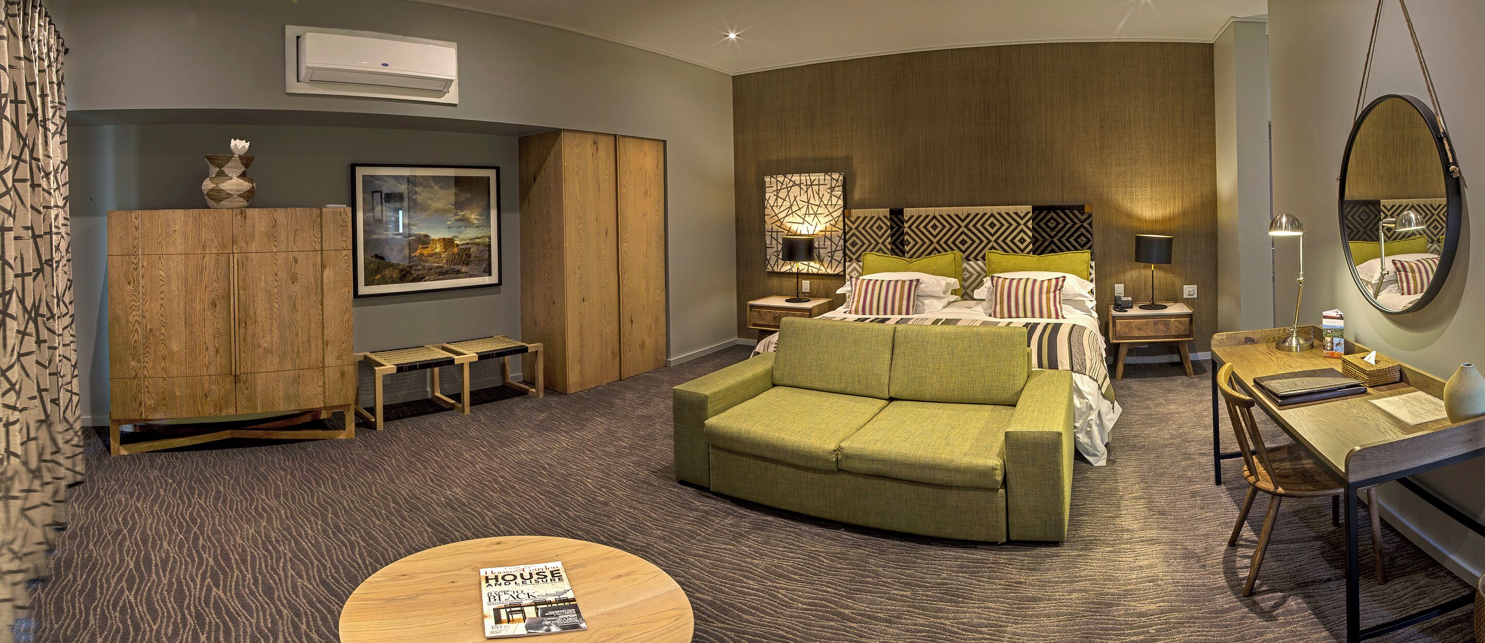 Een moderne hotelkamer met een groene bank, een houten bureau, een hangstoel en een groot bed met gestreepte kussens.