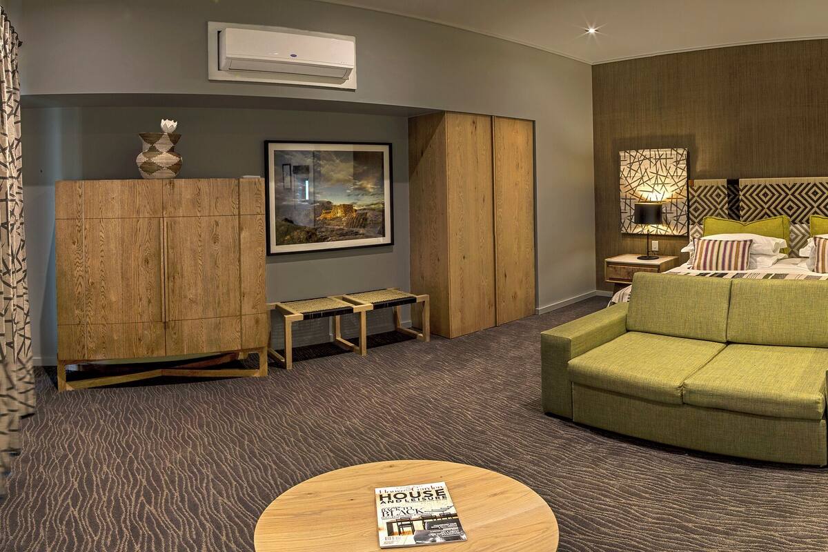 Ein modernes Hotelzimmer mit einem grünen Sofa, einem Holztisch, einem Hängesessel und einem großen Bett mit gemusterten Kissen.