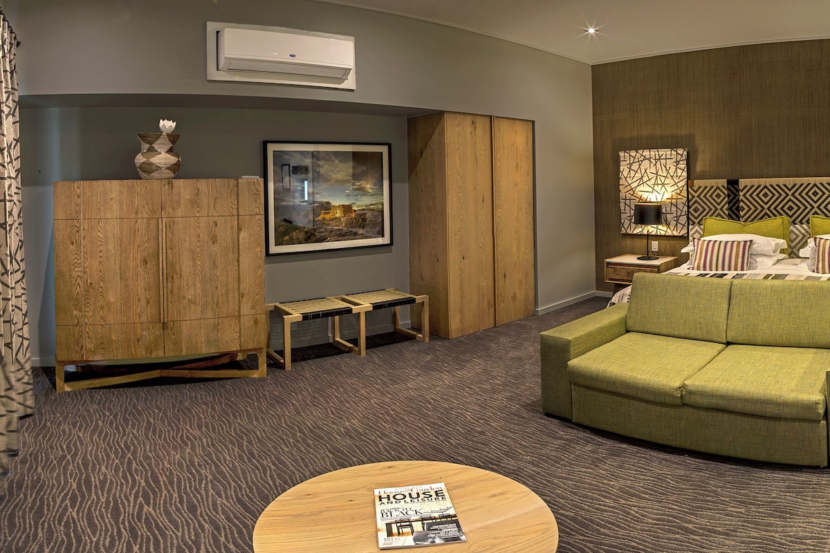 A hotel room with a green sofa, a wooden desk, a hanging chair, and a bed with patterned pillows.