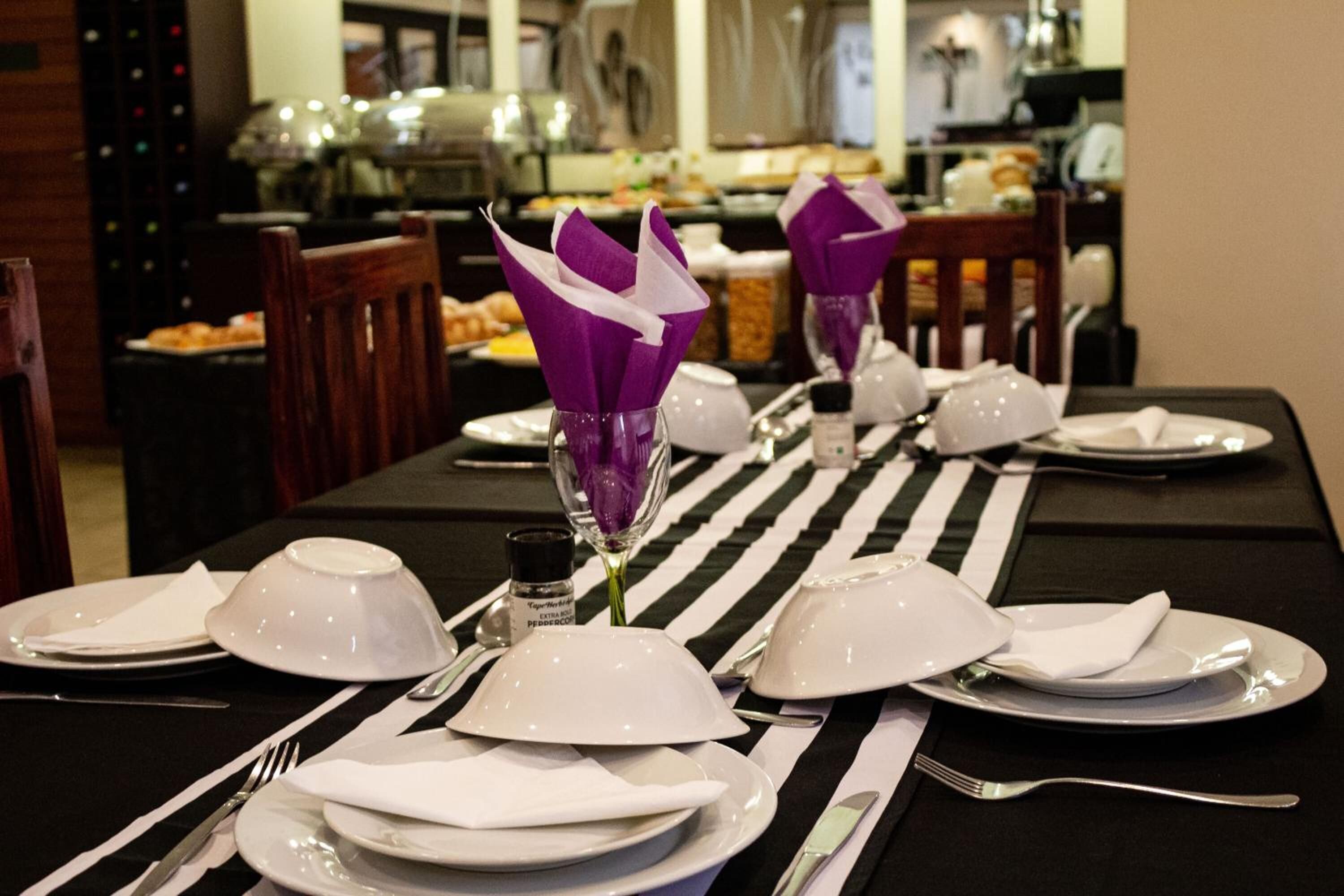 Una mesa de comedor dispuesta con platos, fuentes y cubiertos blancos, adornada con porta servilletas morados y un camino de mesa a rayas.
