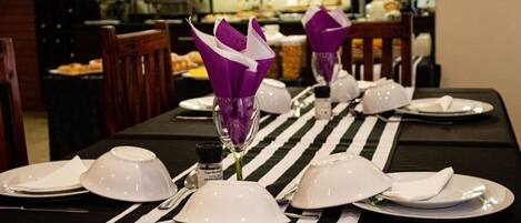 Una mesa de comedor dispuesta con platos, fuentes y cubiertos blancos, adornada con porta servilletas morados y un camino de mesa a rayas.